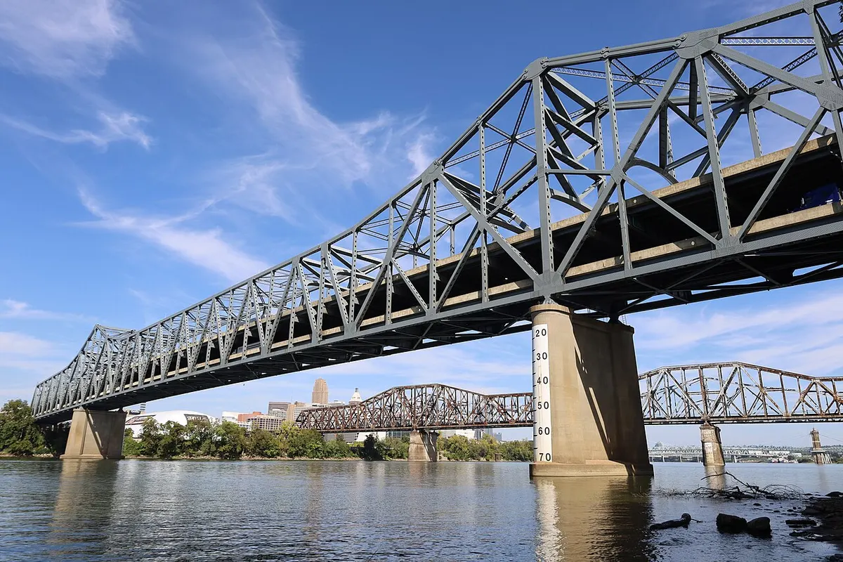 Gray steel double-deck truss bridge on concrete piers crossing the Ohio River, seen from below at water level, with another truss bridge and the Cincinnati skyline behind