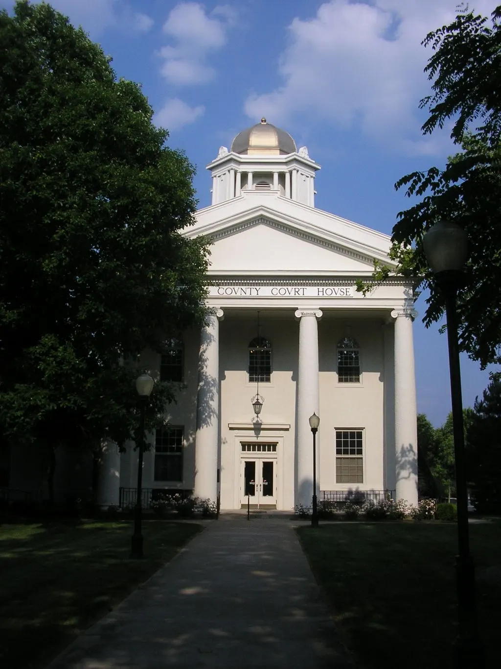 White Greek Revival courthouse with four tall columns, a pediment and a domed cupola, seen at the end of a shaded front walk between trees