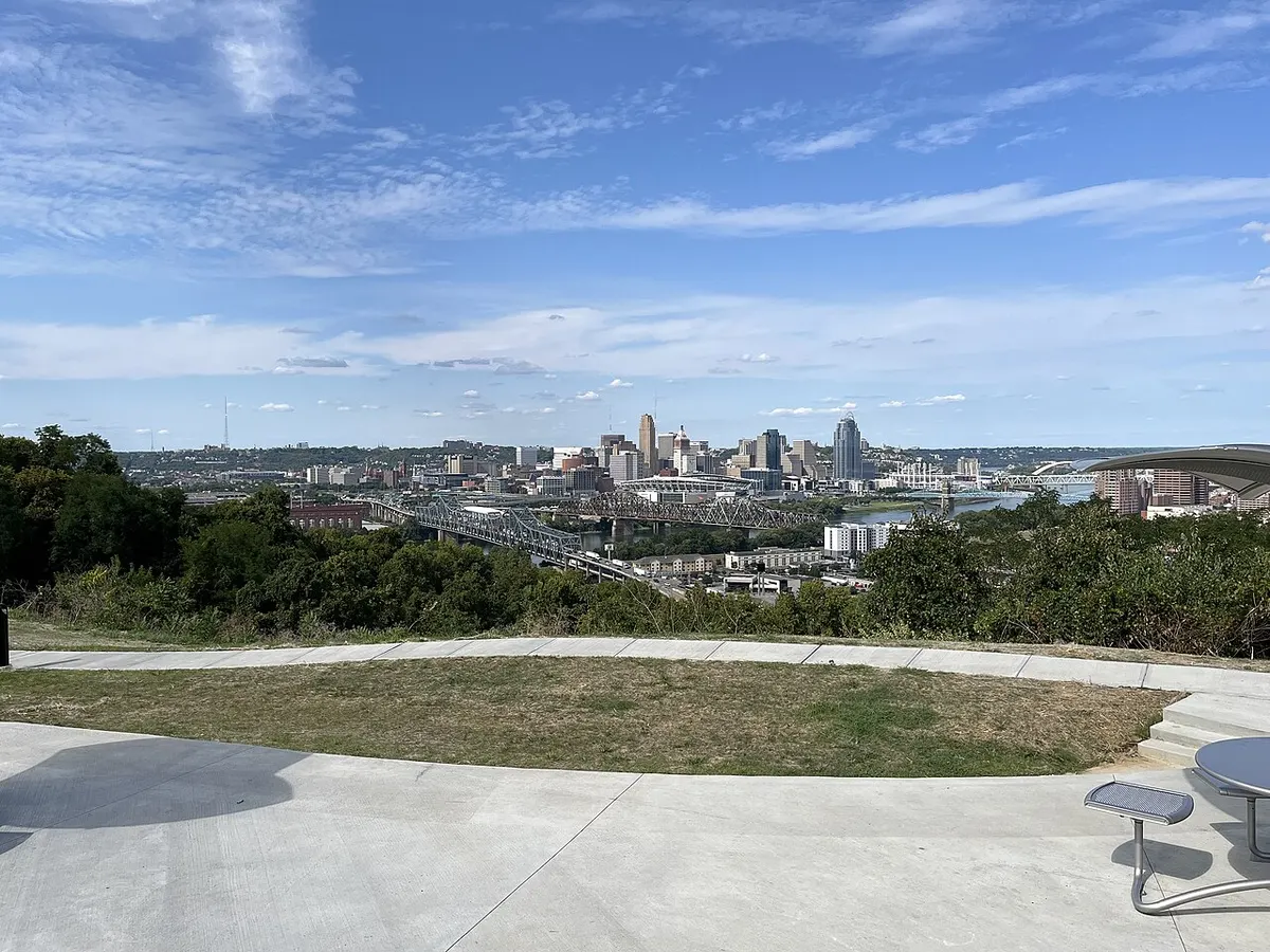 Paved hilltop overlook with a lawn and picnic table, looking over trees toward the Ohio River bridges and the downtown Cincinnati skyline under a blue sky