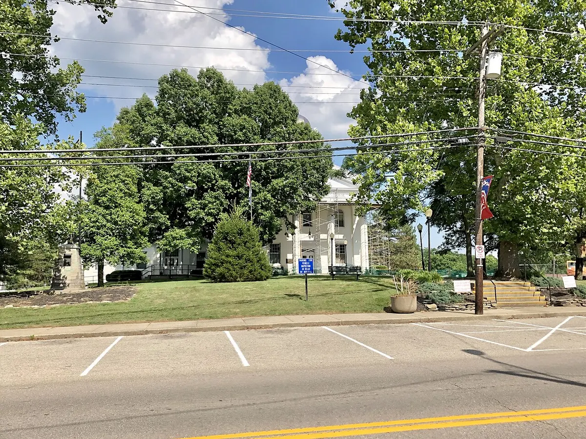 Street view of the white Kenton County Courthouse partly hidden by large trees and scaffolding, with a blue sign on the lawn reading "Parking for vehicle inspection in rear of building" and empty parking spaces in the foreground