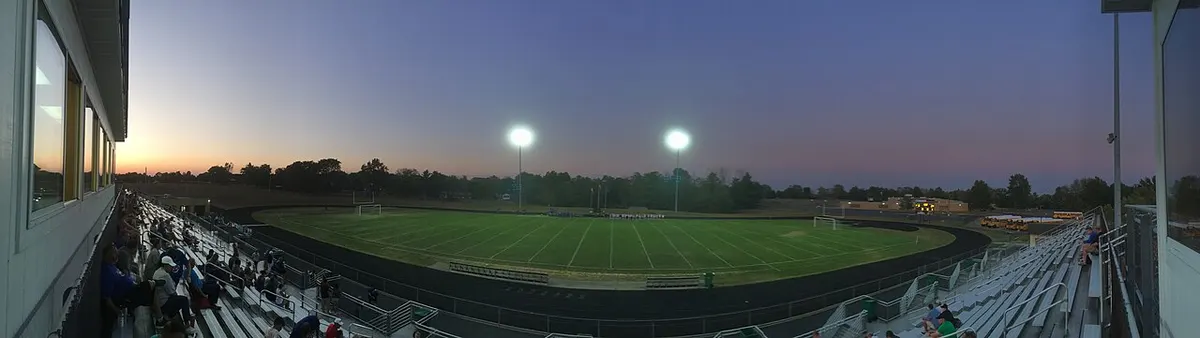 Panoramic view at dusk from the bleachers of a lit football stadium, with spectators in the stands and a tree line under a purple sky