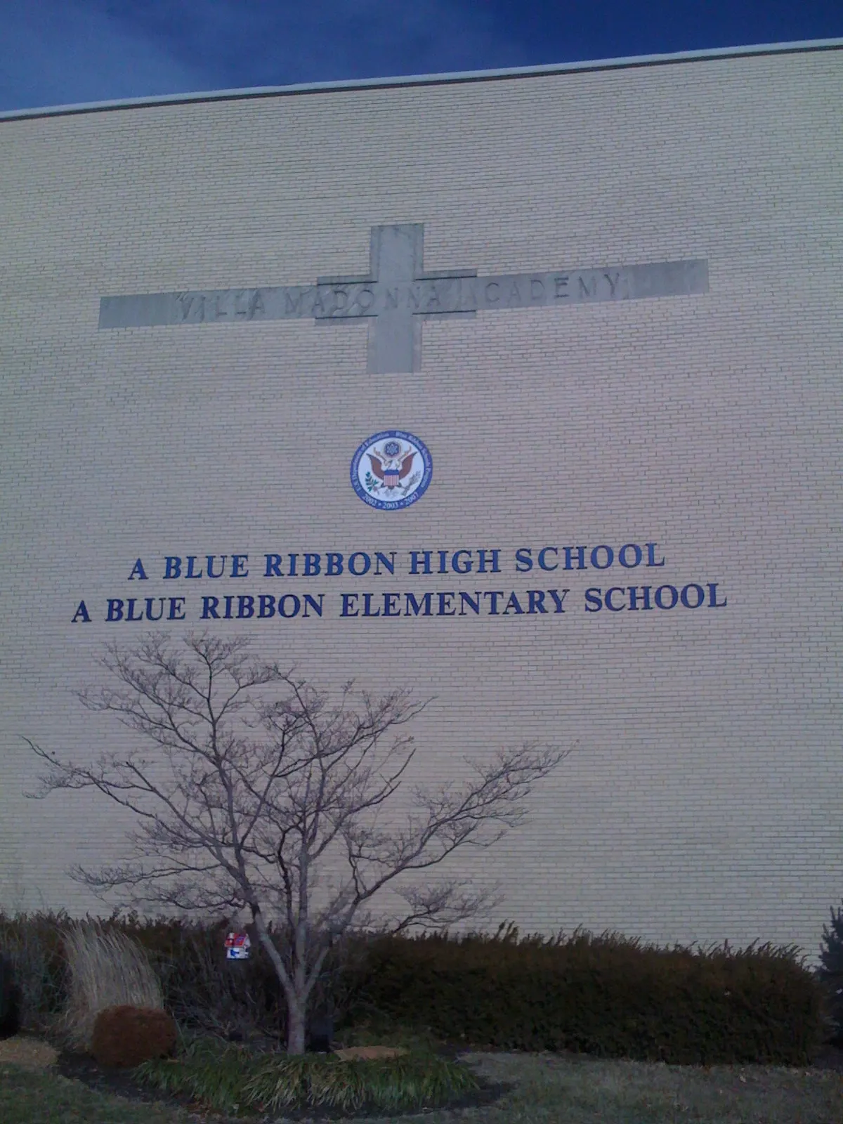 Tan brick wall of Villa Madonna Academy with a stone cross carved with the school name, a U.S. Department of Education seal and lettering reading "A Blue Ribbon High School, A Blue Ribbon Elementary School", above a small bare tree and shrubs